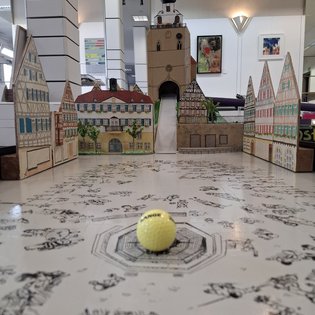 Minigolfbahn mit Modellhäusern und einem gelben Ball im Vordergrund in einem Innenraum