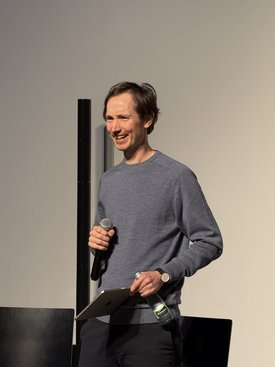 Ein Mann hält ein Mikrofon und eine Wasserflasche bei einer Präsentation beim Symposium "Kunst im Game".