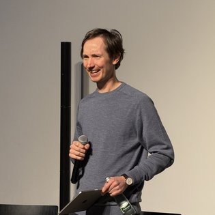 Ein Mann hält ein Mikrofon und eine Wasserflasche bei einer Präsentation beim Symposium "Kunst im Game".