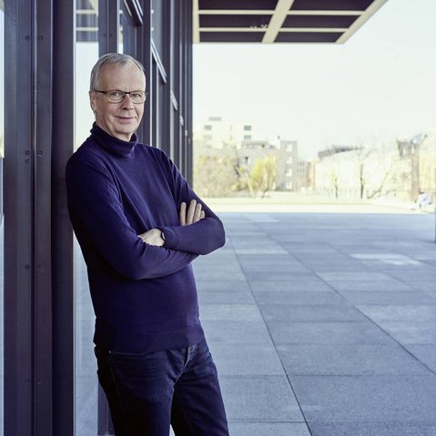 Christoph Biermann mit verschränkten Armen lehnt an Glasfassade eines modernen Gebäudes, trägt dunklen Pullover und Jeans.