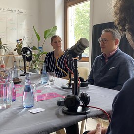 Während der Podcast-Aufnahme: Michael Rubinstein und Bettina Pau sitzen an einem Tisch und haben jeweils ein Mikrofon vor sich stehen. Am Rande der Aufnahme ist noch die Silhouette von Dr. Robert Ogman zu erkennen – ebenfalls mit einem Mikrofon.