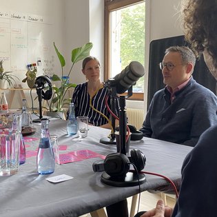 Während der Podcast-Aufnahme: Michael Rubinstein und Bettina Pau sitzen an einem Tisch und haben jeweils ein Mikrofon vor sich stehen. Am Rande der Aufnahme ist noch die Silhouette von Dr. Robert Ogman zu erkennen – ebenfalls mit einem Mikrofon.