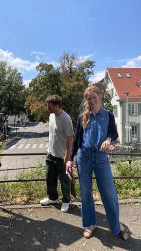 Zwei Personen stehen auf einem Gehweg vor einer Straße mit Zebrastreifen, im Hintergrund Häuser und Bäume, die Frau trägt blaue Kleidung und lächelt, der Mann schaut zur Seite.