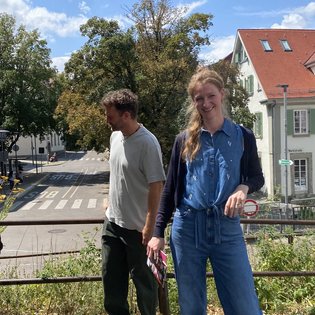 Zwei Personen stehen auf einem Gehweg vor einer Straße mit Zebrastreifen, im Hintergrund Häuser und Bäume, die Frau trägt blaue Kleidung und lächelt, der Mann schaut zur Seite.
