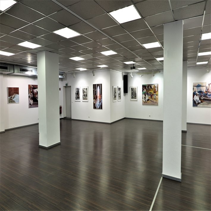 Innenansicht einer modernen Kunstgalerie mit weiß gestrichenen Wänden, dunklem Holzboden und Rasterdeckenbeleuchtung. An den Wänden hängen verschiedene Gemälde und Fotografien, die Szenen mit Menschen darstellen. Die Galerie ist geräumig, gut beleuchtet und wirkt ruhig und einladend.
