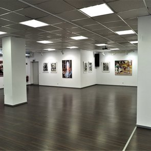 Innenansicht einer modernen Kunstgalerie mit weiß gestrichenen Wänden, dunklem Holzboden und Rasterdeckenbeleuchtung. An den Wänden hängen verschiedene Gemälde und Fotografien, die Szenen mit Menschen darstellen. Die Galerie ist geräumig, gut beleuchtet und wirkt ruhig und einladend.