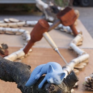 Blaues Katzenmodell liegt auf einem Ast vor einem Minigolf-Parcours aus Holz und Naturmaterialien