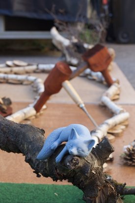 Blaues Katzenmodell liegt auf einem Ast vor einem Minigolf-Parcours aus Holz und Naturmaterialien