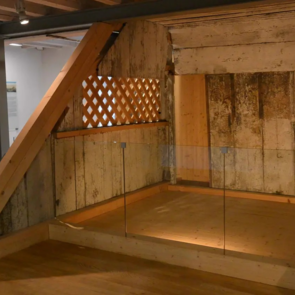 Holzkonstruktion einer historischen Frauenempore aus einer Synagoge, ausgestellt in einem modernen Museumsraum. Die Empore besteht aus gealtertem, weißlich verwittertem Holz mit einer Holzgitteröffnung und wird teilweise von Glaswänden umrahmt. Im Hintergrund befinden sich Informationstafeln und architektonische Modelle.