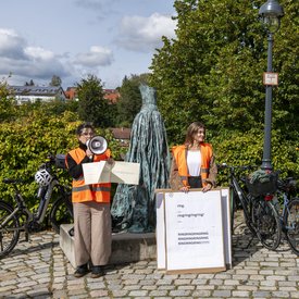 Probe für Mauricio Kagels Stück »Eine Brise – Flüchtige Aktion für 111 Radfahrer«. Eine weiblich gelesene Person mit Warnweste spricht in ein Megafon und hält die Partitur des Stücks in der Hand. Eine weitere weiblich gelesene Person hält mehrere große Plakate in der Hand.