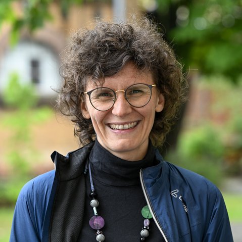 Portrait von Lucia Leidenfrost. Lucia Leidenfrost hat eher kurz geschnittene Locken und trägt eine Brille. Bekleidet ist sie mit einem schwarzen Rollkragenpulli, einer blauen Jacke und einer auffälligen, langen Perlenkette. Sie lächelt offen in die Kamera.