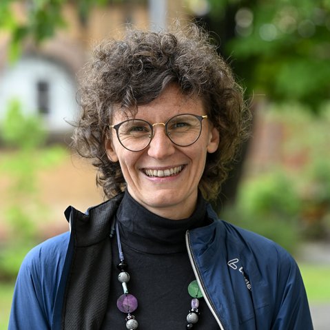 Portrait von Lucia Leidenfrost. Lucia Leidenfrost hat eher kurz geschnittene Locken und trägt eine Brille. Bekleidet ist sie mit einem schwarzen Rollkragenpulli, einer blauen Jacke und einer auffälligen, langen Perlenkette. Sie lächelt offen in die Kamera.