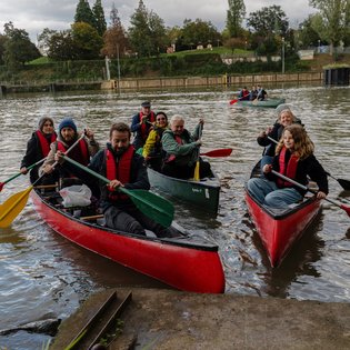 Drei mit Personen besetzte Kanus, die gerade am Ufer anlegen. Alle Personen tragen Schwimmwesten und halten ein Paddel in den Händen.