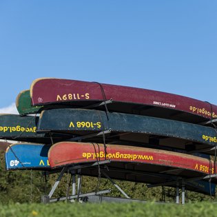Anhänger, der mit Kanus in verschiedenen Farben beladen ist. Der Anhänger steht auf einer Wiese. Der Himmel ist blau.