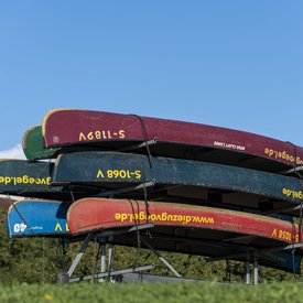Anhänger, der mit Kanus in verschiedenen Farben beladen ist. Der Anhänger steht auf einer Wiese. Der Himmel ist blau.