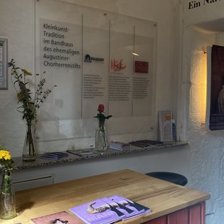Wand mit Texttafeln zur Kleinkunst-Tradition im Bandhaus, davor ein Holztresen mit Magazinen und Vasen mit Blumen, rechts ein beleuchtetes Bild an der Wand.