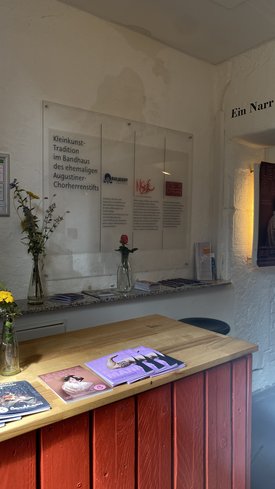 Wand mit Texttafeln zur Kleinkunst-Tradition im Bandhaus, davor ein Holztresen mit Magazinen und Vasen mit Blumen, rechts ein beleuchtetes Bild an der Wand.