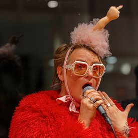 Person mit rotem, flauschigem Oberteil und rosa Haarschmuck mit ausgestrecktem Mittelfinger hält Mikrofon und singt