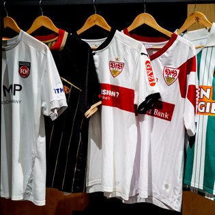 Mehrere Fußballtrikots hängen an Holzbügeln, darunter weiße mit roten Querstreifen und Vereinswappen, ein schwarzes mit goldenen Streifen und ein weißes mit Vereinslogo.