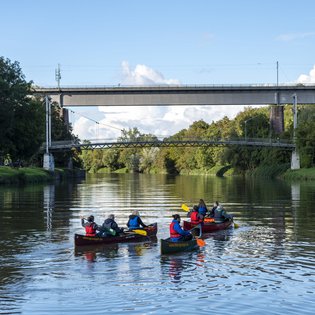 Drei mit Personen besetzte Kanus, die nah aneinander auf einem Gewässer fahren. Das Ufer ist bewaldet. Im Hintergrund ist eine Fußgänger- und Eisenbahnbrücke zu erkennen.