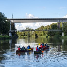 Drei mit Personen besetzte Kanus, die nah aneinander auf einem Gewässer fahren. Das Ufer ist bewaldet. Im Hintergrund ist eine Fußgänger- und Eisenbahnbrücke zu erkennen.