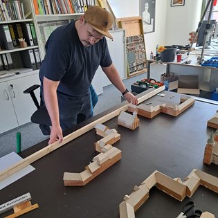 Mann mit brauner Kappe steht an Tisch mit Holzmodell eines Gebäudekomplexes in einem Büro mit Regalen und Werkzeugen