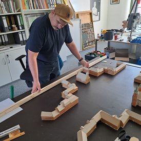 Mann mit brauner Kappe steht an Tisch mit Holzmodell eines Gebäudekomplexes in einem Büro mit Regalen und Werkzeugen
