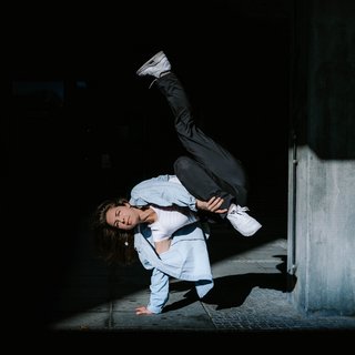 Sophie M. Lindner in einer Breaking-Position, aufgestützt auf nur einem Arm. Rechts neben ihr ein Beton-Element. Der Hintergrund des Bildes ist sehr dunkel bis schwarz gehalten.