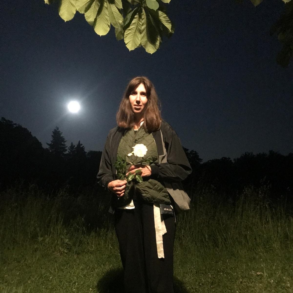 Portrait von Gloria Aino Grzywatz bei Nacht. Sie hat langes, braunes Haar, trägt zwei Jacken übereinander und hat eine Tasche über die Schulter gehängt. In der Hand hält sie eine einzelne weiße Blume.