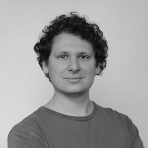 Porträt von Chris Binder. Er hat lockige, dunkle Haare. Er trägt ein schlichtes, dunkles T-Shirt und blickt ruhig und direkt in die Kamera. Der Hintergrund ist neutral und hell, was den Fokus auf das Gesicht legt. Das Bild ist in Schwarz-Weiß gehalten.