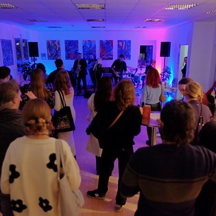 Bild einer Vernissage in den Räumlichkeiten des PLEKS. Alles ist in bläuliches Licht getaucht. An den Wänden hängen Gemälde, im hinteren Teil des Raumes spielt eine Band, im vorderen Teil stehen Menschen, die der Band zuhören.