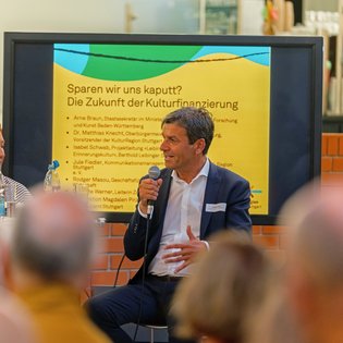 Rodger Masou spricht ins Mikrofon, im Hintergrund ein Bildschirm der den Titel zeigt: Sparen wir uns kaputt? Die Zukunft der Kulturfinanzierung