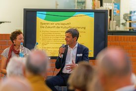 Rodger Masou spricht ins Mikrofon, im Hintergrund ein Bildschirm der den Titel zeigt: Sparen wir uns kaputt? Die Zukunft der Kulturfinanzierung