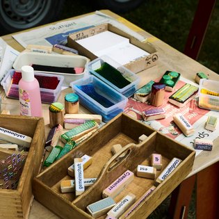 Nahaufnahme von bunten Stempeln und Farben auf einem Holztisch. Auf den Stempeln stehen Begriffe in verschiedenen Sprachen wie: Gemeinschaft, Utopie, Vielfalt.