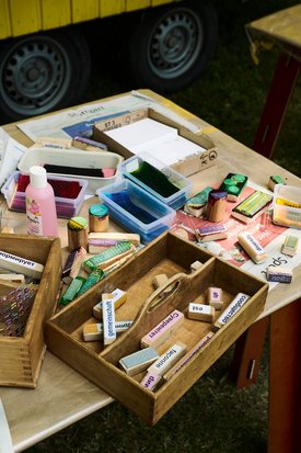 Nahaufnahme von bunten Stempeln und Farben auf einem Holztisch. Auf den Stempeln stehen Begriffe in verschiedenen Sprachen wie: Gemeinschaft, Utopie, Vielfalt.