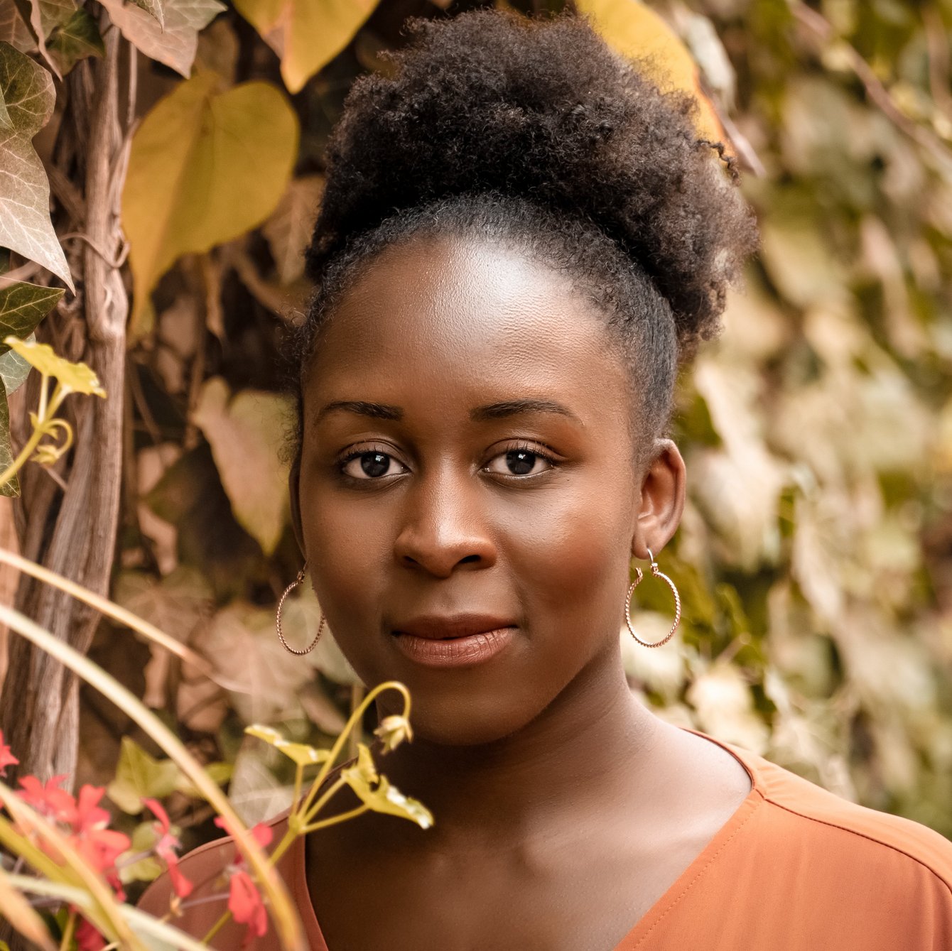 Portrait von Yeama Bangali, aufgenommen vor einer Wand aus Efeu. Sie hat lockiges, schwarzes Haar, das zu einem Zopf zusammengebunden ist. Sie trägt goldene Creolen und ein braunes Shirt.