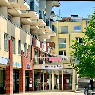 Außenansicht der Städtischen Galerie in Filderstadt an einem sonnigen Tag. Das Galeriegebäude befindet sich im Erdgeschoss eines modernen Wohn- und Geschäftshauses mit rot-beiger Fassade und mehreren Balkonen. Vor dem Eingang mit Glasfront und Beschriftung ‚städtische galerie‘ stehen Sitzbänke und ein grüner Baum spendet Schatten. Eine Treppe führt rechts vom Gebäude hinauf.