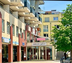 Außenansicht der Städtischen Galerie in Filderstadt an einem sonnigen Tag. Das Galeriegebäude befindet sich im Erdgeschoss eines modernen Wohn- und Geschäftshauses mit rot-beiger Fassade und mehreren Balkonen. Vor dem Eingang mit Glasfront und Beschriftung ‚städtische galerie‘ stehen Sitzbänke und ein grüner Baum spendet Schatten. Eine Treppe führt rechts vom Gebäude hinauf.