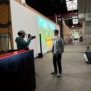 Ein Mann spricht auf einem Symposium über Kunst im Spiel, während ein anderer Mann ihn filmt.