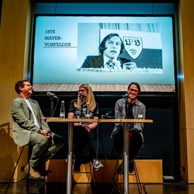 Drei Personen sitzen an einem Tisch mit Mikrofonen, im Hintergrund eine Leinwand mit einem schwarz-weißen Foto und dem Text '1975 MAYER-VORFELDER'.