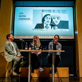 Drei Personen sitzen an einem Tisch mit Mikrofonen, im Hintergrund eine Leinwand mit einem schwarz-weißen Foto und dem Text '1975 MAYER-VORFELDER'.