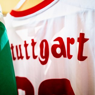 Weißes Trikot mit roter Aufschrift 'stuttgart' und rotem Kragenrand, teilweise verdeckt von grünem Stoff.