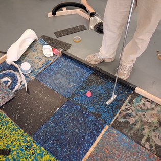 Person steht auf einem Boden mit bunten Teppichfliesen und hält einen Golfschläger, ein pinkfarbener Ball liegt auf dem Boden