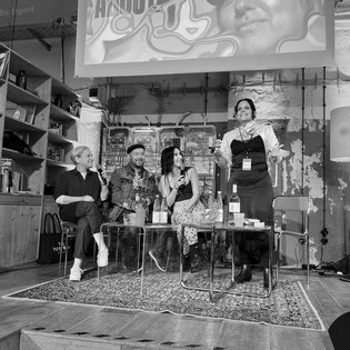 Die drei Gäst*innen sitzen in einer Reihe auf Stühlen auf der Bühne. Mirna Funk und Vivian Kanner halten jeweils ein Mikro in der Hand. Moderatorin Anna Lampert hat sich von ihrem Stuhl erhoben und scheint Worte an das Publikum zu richten. Alle 4 Personen auf dem Bild lachen herzlich.
