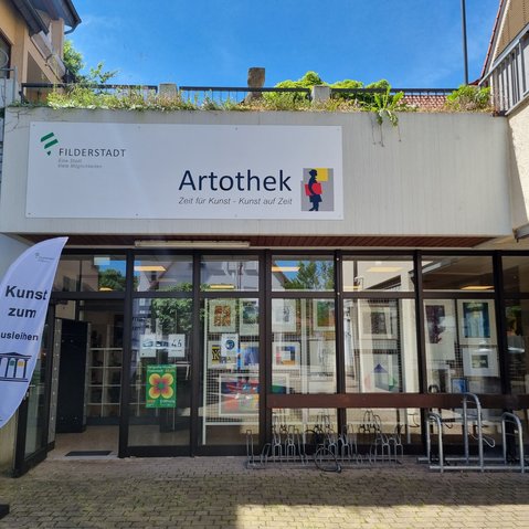 Eingangsbereich der Artothek Filderstadt bei sonnigem Wetter. Über dem Eingang hängt ein großes Schild mit dem Logo der Stadt Filderstadt und dem Schriftzug „Artothek – Zeit für Kunst – Kunst auf Zeit“. Links neben der Tür steht eine weiße Fahne mit der Aufschrift „Kunst zum Ausleihen“. Durch die Glasfront sind zahlreiche gerahmte Kunstwerke im Innenraum sichtbar. Vor dem Eingang befinden sich Fahrradständer.