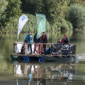 Das Festival-Floß unterwegs auf einem Gewässer in der Region. An Bord sind verschiedene Personen. Eine Person schwingt eine Fahne. Ein Banner an der Reling des Floßes zeigt das Logo von JETZT!.