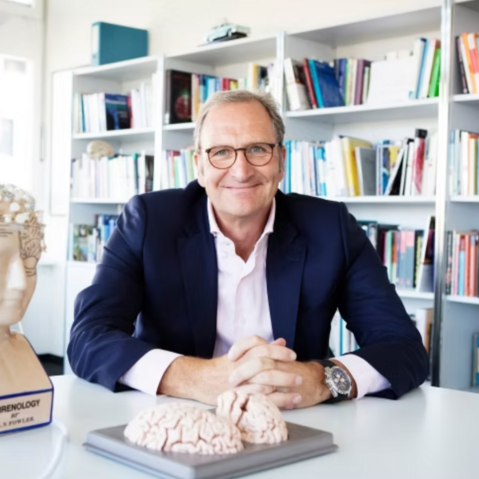 Dr. h.c. Lutz Jäncke sitzt an einem weißen Tisch und hat die Hände auf dem Tisch gefaltet. Er trägt eine Brille und ist mit einem weißen Hemd und ein blauen Jacket bekleidet. Vor ihm und neben ihm auf dem Tische stehen Modelle des Gehirns. Im Hintergrund ist ein großes Bücherregal zu sehen.