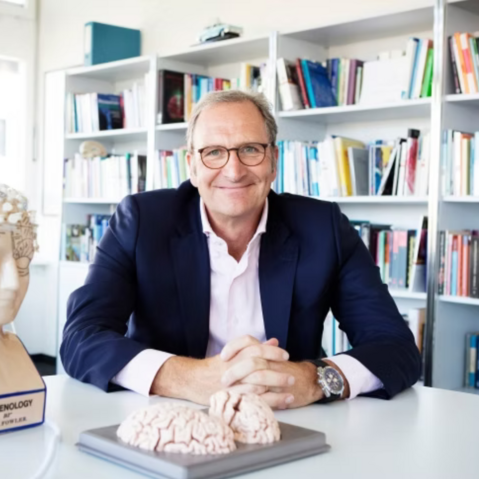 Dr. h.c. Lutz Jäncke sitzt an einem weißen Tisch und hat die Hände auf dem Tisch gefaltet. Er trägt eine Brille und ist mit einem weißen Hemd und ein blauen Jacket bekleidet. Vor ihm und neben ihm auf dem Tische stehen Modelle des Gehirns. Im Hintergrund ist ein großes Bücherregal zu sehen.