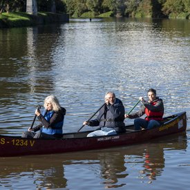 Ein mit drei Personen besetztes Kanu, das auf einem Gewässer fährt. Alle drei Personen tragen Schwimmwesten und halten ein Paddel in den Händen.