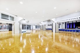 Innenansicht auf das Foyer und den unbestuhlten Kombisaal. Die Wände sind in hellen Tönen gehalten, der Saal verfügt über einen Parkettboden. Hinten im Saal ist eine Bühne zu sehen